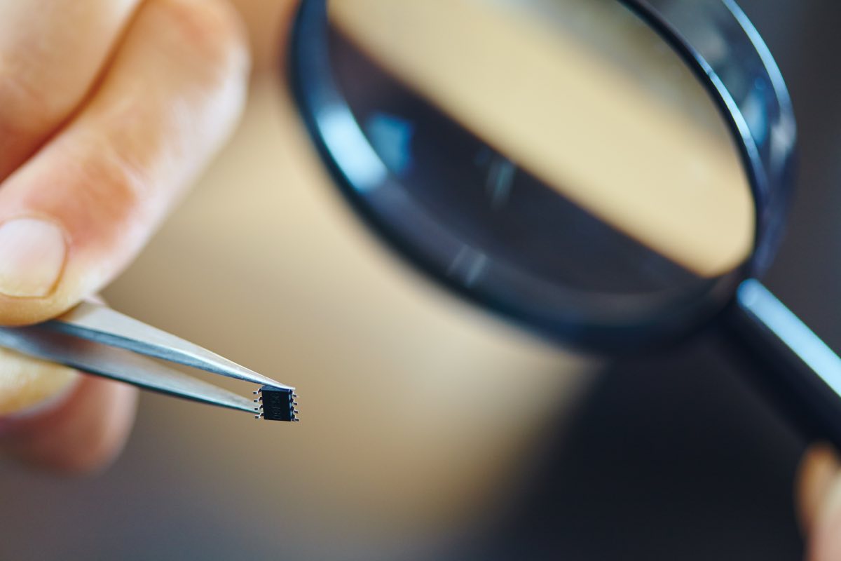 A surface-mount component held in tweezers being checked under a magnifier
